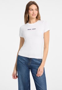 Valkoinen puuvillapaita, jossa on istuva malli ja lyhyet hihat. Edessä keskustassa on sininen ja punainen "Guess Jeans" -logo.