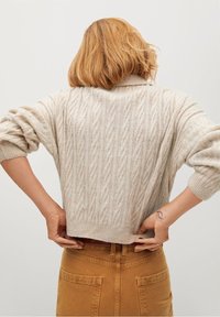 Pull en maille torsadée de couleur crème avec une coupe décontractée et des épaules tombantes, doté d'un col haut et d'un ourlet côtelé, associé à un jean marron taille haute.