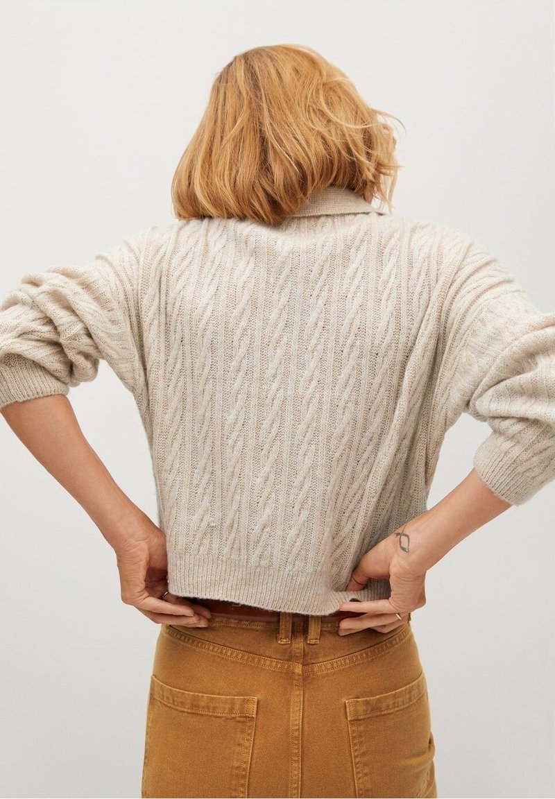 Pull en maille torsadée de couleur crème avec une coupe décontractée et des épaules tombantes, doté d'un col haut et d'un ourlet côtelé, associé à un jean marron taille haute.