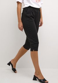 Czarne, krótke spodnie z plisami, tapered fit oraz detalem w postaci rozcięcia u dołu, w połączeniu z sandałami na obcasie typu block heel odkrytymi na palcach. Gładka faktura materiału.