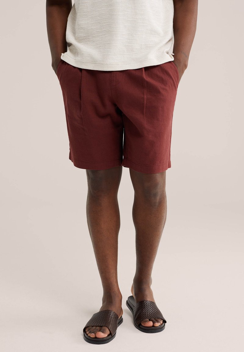 Homme portant un short couleur rouille, une chemise blanche texturée et des sandales tressées marron foncé, debout les mains dans les poches.