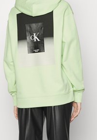 Persoon draagt een lichtgroene hoodie met een zwart-wit Calvin Klein Jeans logo op de achterkant en zwarte leren broek.