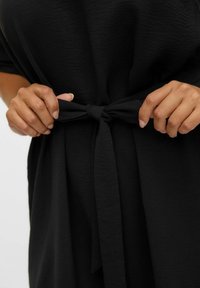 Zwarte stoffen jurk met een getextureerde uitstraling, voorzien van een strik op de taille van hetzelfde materiaal, wat zorgt voor een getailleerd silhouet.