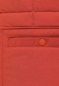 Κόκκινο επενδυμένο ύφασμα με ορθογώνια τσέπη και αντίστοιχο πορτοκαλ ί κουμπί με πατητό. Υφή με ορατές ραφές.