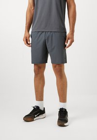 Mees, kellel on seljas tumehallid treeningshortsid, hall särk, valged sokid ja mustad Nike jooksujalatsid, seisab valgel taustal.