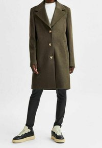 Cappotto di lana verde oliva con colletto a revers, tre bottoni e tasche laterali, abbinato a pantaloni neri aderenti e sneaker nere con dettagli crema.