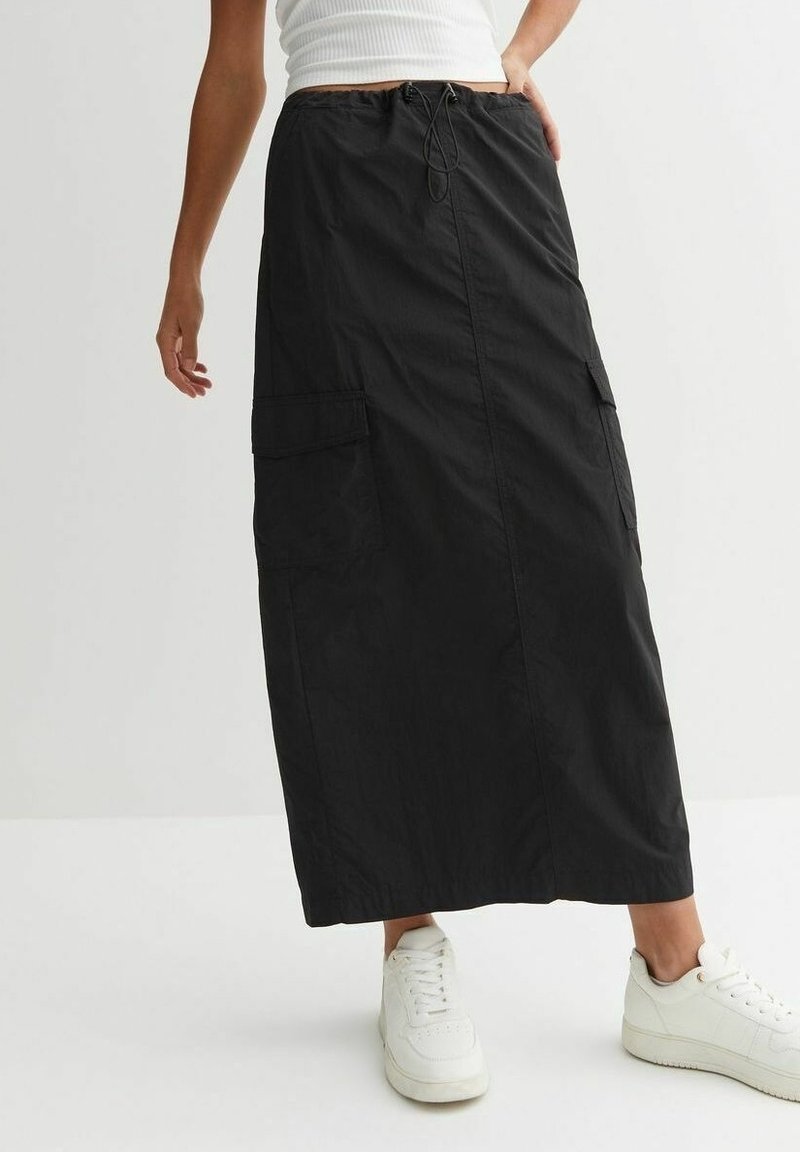 Vrouw in een zwarte lange cargoskirt met zijzakken, witte sneakers en een witte mouwloze top tegen een effen achtergrond.