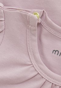 Ljusrosa bomullsplagg med rund halsringning, med en volangkant och en gyllene tryckknappdetalj på axeln. Mjuk textur.