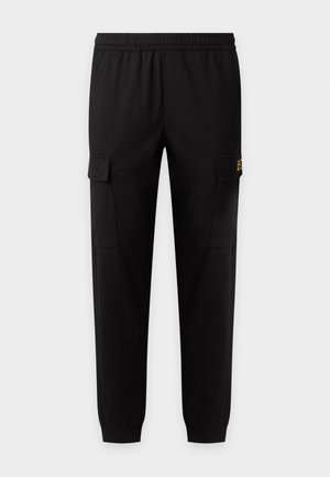 Joven con sudadera gris con cremallera, camiseta blanca, pantalones cargo negros, zapatillas blancas y gorra de béisbol negra, de pie frente a un fondo liso.