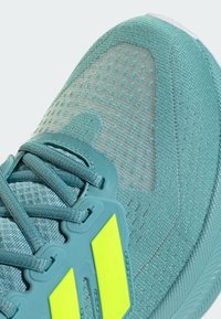 Zapatilla deportiva de malla aqua con una parte superior texturizada, acentos amarillos prominentes, cordones asegurados y un diseño de suela flexible.