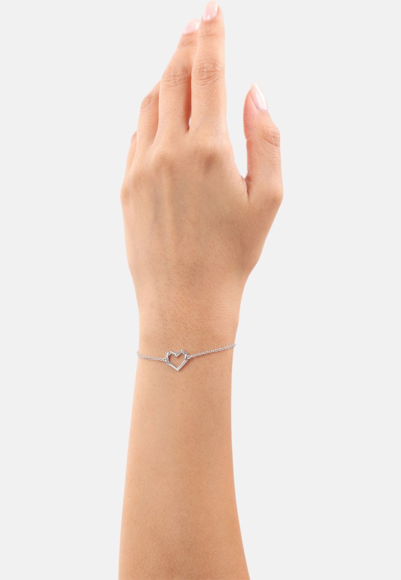 Bracciale d'argento con un ciondolo a forma di cuore su una delicata catena, appoggiato su un polso di tonalità della pelle. Design semplice con minimi accenti.