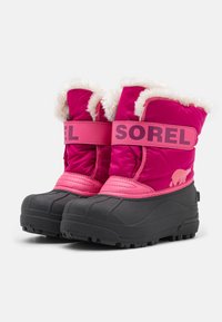 Stivali da neve isolati rosa con rivestimento bianco soffice, suole nere in gomma e una larga fascia rosa con la scritta "SOREL" sulla parte anteriore.