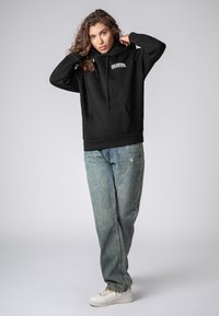 Schwarzer Kapuzenpullover mit einer Fronttasche, mit weißem Textlogo; kombiniert mit ausgewaschenen blauen Jeans und weißen Sneakers.