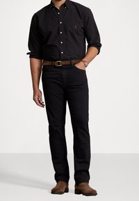 Svart skjorta med knappar och en liten logotyp, svarta byxor, brunt bälte med ett guldspänne och bruna mockastövlar. Klassisk, åtsittande design.