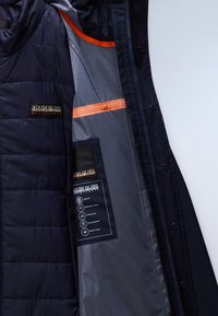 Marineblaue gesteppte Jacke mit orangenem Besatz, geöffnet, um das Innenetikett mit Angaben zu modularen, atmungsaktiven, abgeklebt genähten Nähten, wasserabweisendem Ripstop-Stoff zu zeigen.