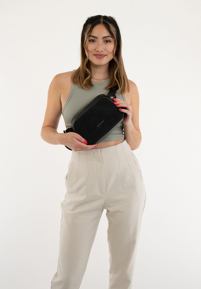 Borsa a tracolla nera in suede con accessori dorati, forma rettangolare, texture liscia; modello che indossa un top verde aderente e pantaloni beige.