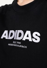 Sweat-shirt noir avec grand texte blanc brodé "ADIDAS", avec les textes plus petits "EST. 1949" et "HERZOGENAURACH" en dessous. Design classique à col rond.