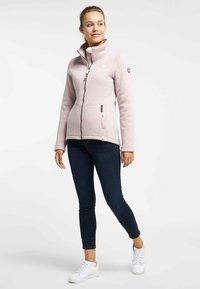 Giacca in pile rosa chiaro con zip e colletto alto, due tasche frontali e logo sulla manica, abbinata a jeans scuri e sneakers bianche.