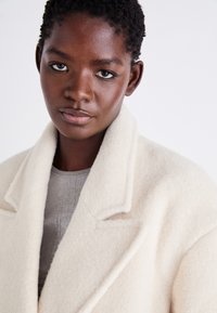 Manteau oversize en mélange de laine crème avec de larges revers, finition texturée et poches profondes, superposé sur un pull gris clair.