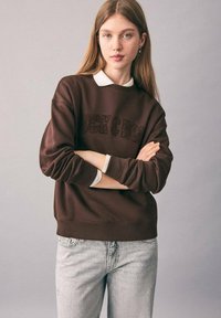 Giovane donna con capelli lunghi che indossa un felpa marrone sovrapposta a una camicia bianca con colletto e jeans chiari, in piedi con le braccia incrociate.