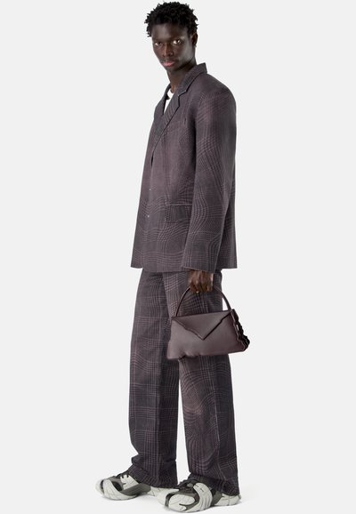 Homme portant un costume à carreaux foncés et des baskets, tenant un sac à main en cuir brun foncé sur un fond uni.