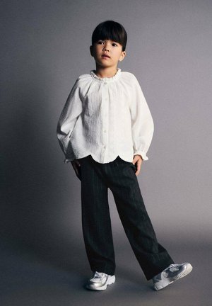 Chemise blanche texturée à boutons, avec un ourlet festonné ; portée avec un pantalon noir à rayures fines et des baskets argentées.