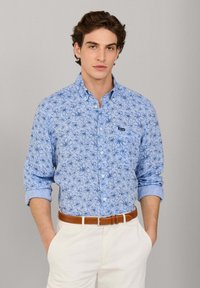Junger Mann, der ein blaues, mit Blumenmuster bedrucktes Hemd mit hochgekrempelten Ärmeln, beigen Hosen und einen braunen Ledergürtel trägt, steht mit den Händen in den Taschen.