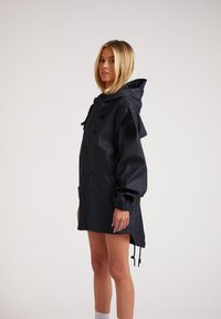Veste imperméable noire avec capuche, manches longues, poignets élastiques et cordon de serrage à l'ourlet. Comprend une poche latérale et un logo cousu sur la manche.