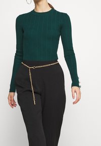 Dunkelgrüner, gerippter Strickpullover mit rundem Hals, kombiniert mit hoch taillierten schwarzen Hosen, die einen goldenen Kettengürtel und Knopfdetails an den Ärmeln aufweisen.