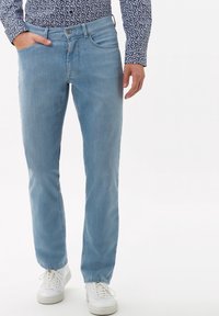 Hombre vistiendo jeans de color azul claro, zapatillas blancas y una camisa de manga larga con patrón, con la mano en el bolsillo, de pie contra un fondo blanco.