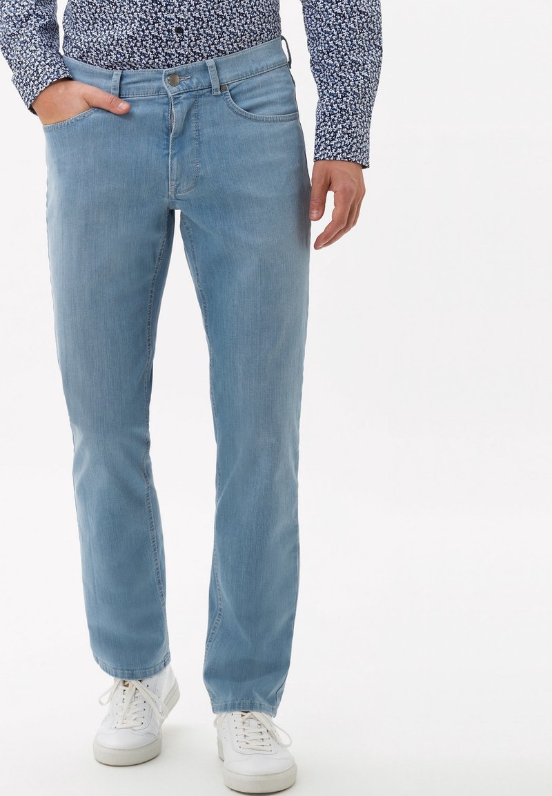 Hombre vistiendo jeans de color azul claro, zapatillas blancas y una camisa de manga larga con patrón, con la mano en el bolsillo, de pie contra un fondo blanco.