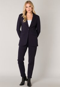 Marineblauw op maat gemaakte pak met een blazer met één knoop, notch revers en zijzakken; gecombineerd met rechte broekspijpen en zwarte enkelboots.