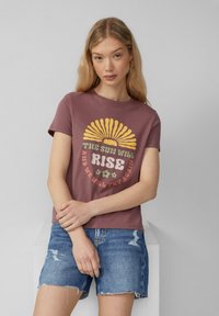T-shirt marron à manches courtes avec un graphique de soleil jaune portant le texte "LE SOLEIL SE RÉVEILLERA" et des accents floraux, associé à un short en denim déchiré.