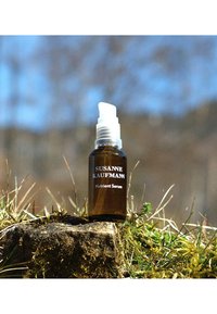 Brązowa szklana butelka z białym dozownikiem. Etykieta z napisem "SUSANNE KAUFMANN" oraz "Nutrient Serum." Ustawiona na kamienistej ziemi z trawą.