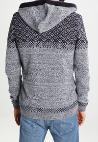 Grau melierter Kapuzenpullover mit strukturiertem Strick. Verfügt über ein dunkles, gemustertes Passe sowie gerippte Bündchen. Die Rückansicht zeigt ein Lagen-Design.