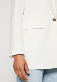 Bílý blazer se strukturovaným designem, s jedním hnědým knoflíkem a přední kapsou. Látka vypadá hladce s matnou texturou.