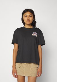 Black cotton t-shirt with short sleeves, round neckline, and colorful graphic print on the left chest. Paired with a beige corduroy skirt.