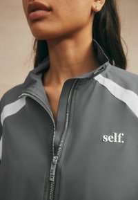 Grey zip-up jacket featuring white accents and a silver logo "self." on the left chest. Constructed from smooth, lightweight fabric.