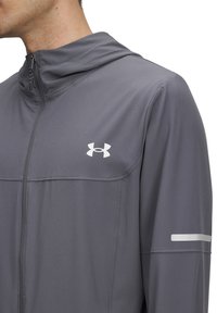 Harmaa urheilu takki, jossa on vetoketjullinen etuosa, huppu ja heijastavat yksityiskohdat hihoissa. Ominaisuudet sileä pinta ja Under Armour -logo.