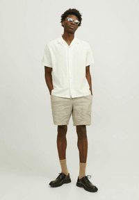 Chemise blanche à manches courtes, short beige, chaussures noires, chaussettes couleur sable. Homme debout les mains dans les poches, portant des lunettes de soleil rondes. Fond blanc uni.