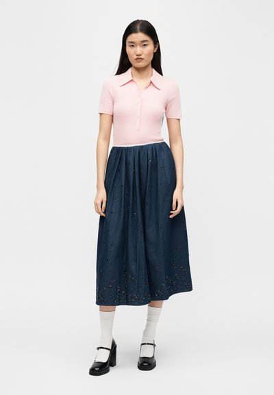 Γυναίκα που στέκεται φορώντας ένα ροζ ανοιχτό ροζ κοντομάνικο πουκάμισο με κουμπιά, φούστα μήκους μίντι σε ναυτικό μπλε με κεντήματα λουλουδιών, λευκές κάλτσες και μαύρα παπούτσια Mary Jane.