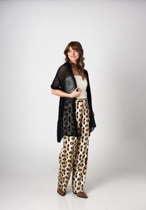 Kobieta stojąca, ubrana w czarny dziergany szal, biały top tubowy, wzorzyste spodnie o szerokich nogawkach i brązowe buty na szpic, na tle jednolitego jasnego tła.