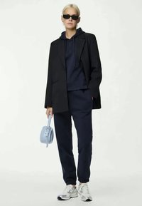 Hoodie azul navy colocado por baixo de um blazer preto, combinado com calças tapered azuis navy. Acessorizado com uma mala azul claro e ténis brancos.