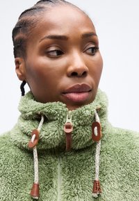 Γυναίκα με πλεξούδες, ντυμένη με ένα ανοιχτοπράσινο πανωφόρι με υφή και ψηλό γιακά, με λευκά κορδόνια, κοιτάζει ελαφρώς προς το πλάι.