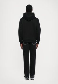 Άνδρας standing με πλάτη, φορώντας μαύρη φούτερ με κουκούλα, μαύρα τζιν και μαύρα-άσπρα αθλητικά παπούτσια σε απλό φόντο.