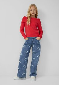 Červený rebrovaný sveter s odhalenými ramenami, skombinovaný so svetlomodrými džínsami s potlačou mašlí. Model má na sebe biele tenisky.