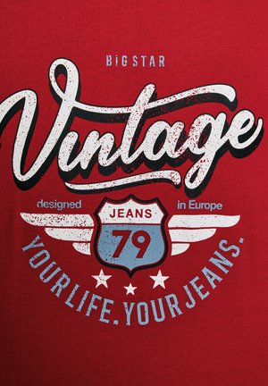 Czerwona koszulka z krótkim rękawem z odważnym, vintage'owym nadrukiem. Charakteryzuje się białym i niebieskim tekstem, wzorem tarczy z napisem "JEANS 79" oraz akcentami w postaci skrzydeł.