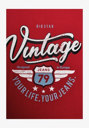 Czerwona koszulka z krótkim rękawem z odważnym, vintage'owym nadrukiem. Charakteryzuje się białym i niebieskim tekstem, wzorem tarczy z napisem "JEANS 79" oraz akcentami w postaci skrzydeł.