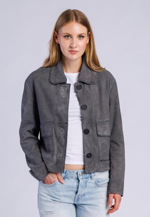 Junge Frau mit langen blonden Haaren, die eine graue Jacke mit Knöpfen über einem weißen Hemd und hellblauen zerrissenen Jeans trägt, steht mit einer Hand in der Tasche.