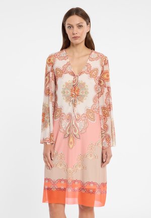 Femme portant une robe à manches longues et longueur genou avec un motif cachemire rose, orange et beige, et un détail noué au niveau de l'encolure.